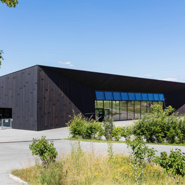 Moderne, schwarze Gebäudearchitektur mit markanter, geneigter Dachform und großflächiger Glasfront. Die Fassade besteht aus dunkel verkohltem Holz (Shou Sugi Ban Optik) und bildet einen starken Kontrast zur umgebenden grünen Landschaft. Im Vordergrund ein Weg, Wildblumen und ein Baum. Am Gebäude ist der Schriftzug „museumstrakt“ erkennbar.