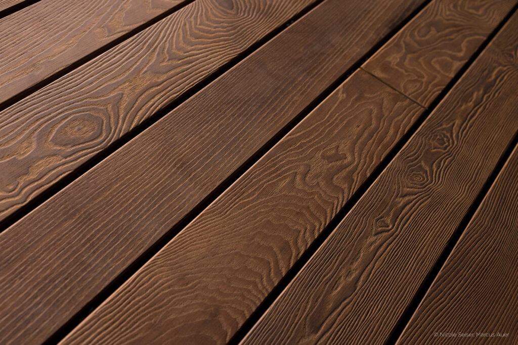 Terrassendielen mit gebürsteter Oberfläche in einem warmen Dunkelbraun. Die ausgeprägte Maserung ist deutlich spürbar und verleiht den Dielen eine lebendige, strukturierte Haptik mit natürlichem, rustikalem Charakter.