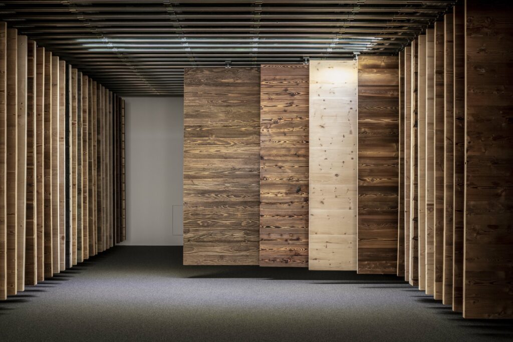 Ein abgedunkelter Raum mit Holzpaneelen in unterschiedlichen Farbtönen, die von der Decke hängen. Der Boden ist mit Teppich ausgelegt, die Decke mit dunklen Lamellen gestaltet – ideal zur Präsentation von Holzoberflächen.