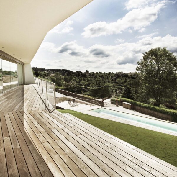 Weitläufige Terrasse aus Mareiner Gardasee Holz mit Glasfront und Ausblick auf Pool und grüne Hügellandschaft. Die klare Linienführung der Architektur kontrastiert mit der natürlichen Umgebung unter teils bewölktem Himmel.